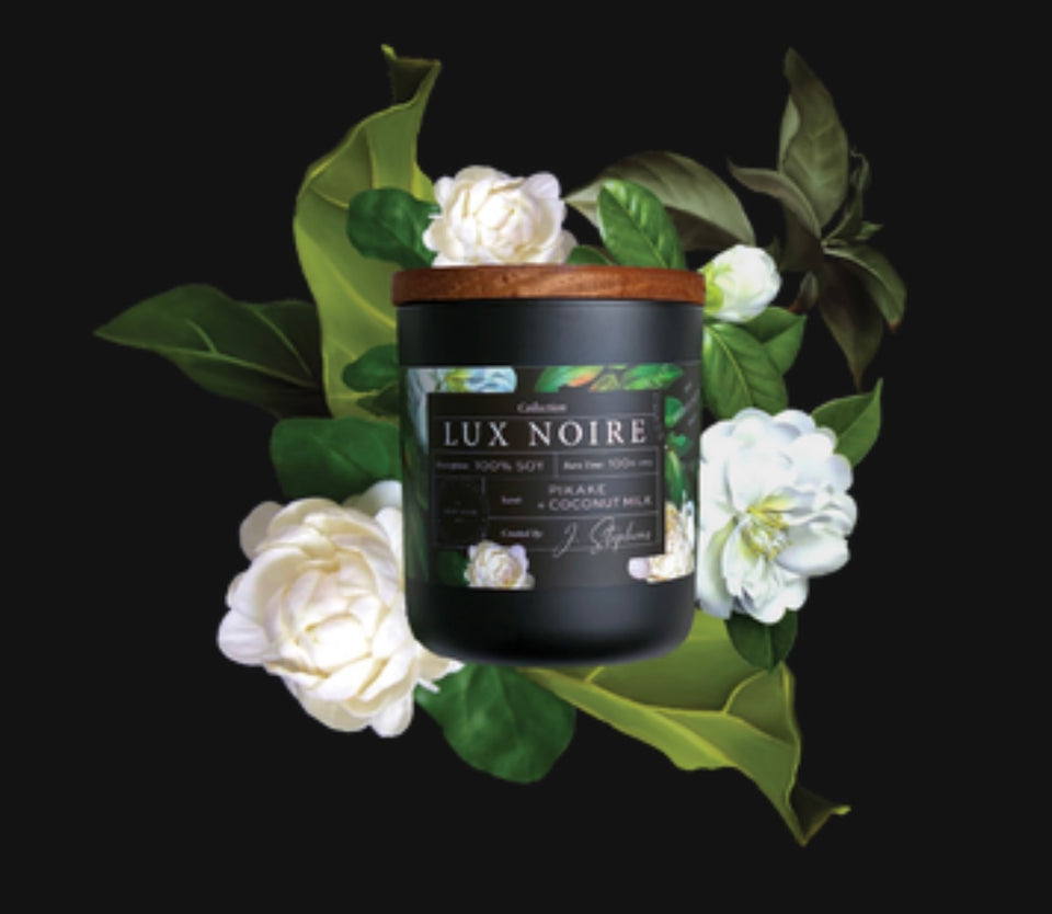 Lux Noire Candle - candle laying on the bed of white flowers.Black frosted glass with wooden top and flowers printed on the label with white Pikake flowers.