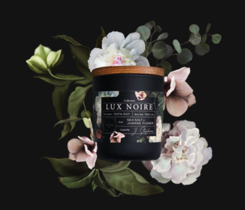 Lux Noire Candle - candle laying on the bed of white and pale pink flowers.Black frosted glass with wooden top and flowers printed on the label with flowers.