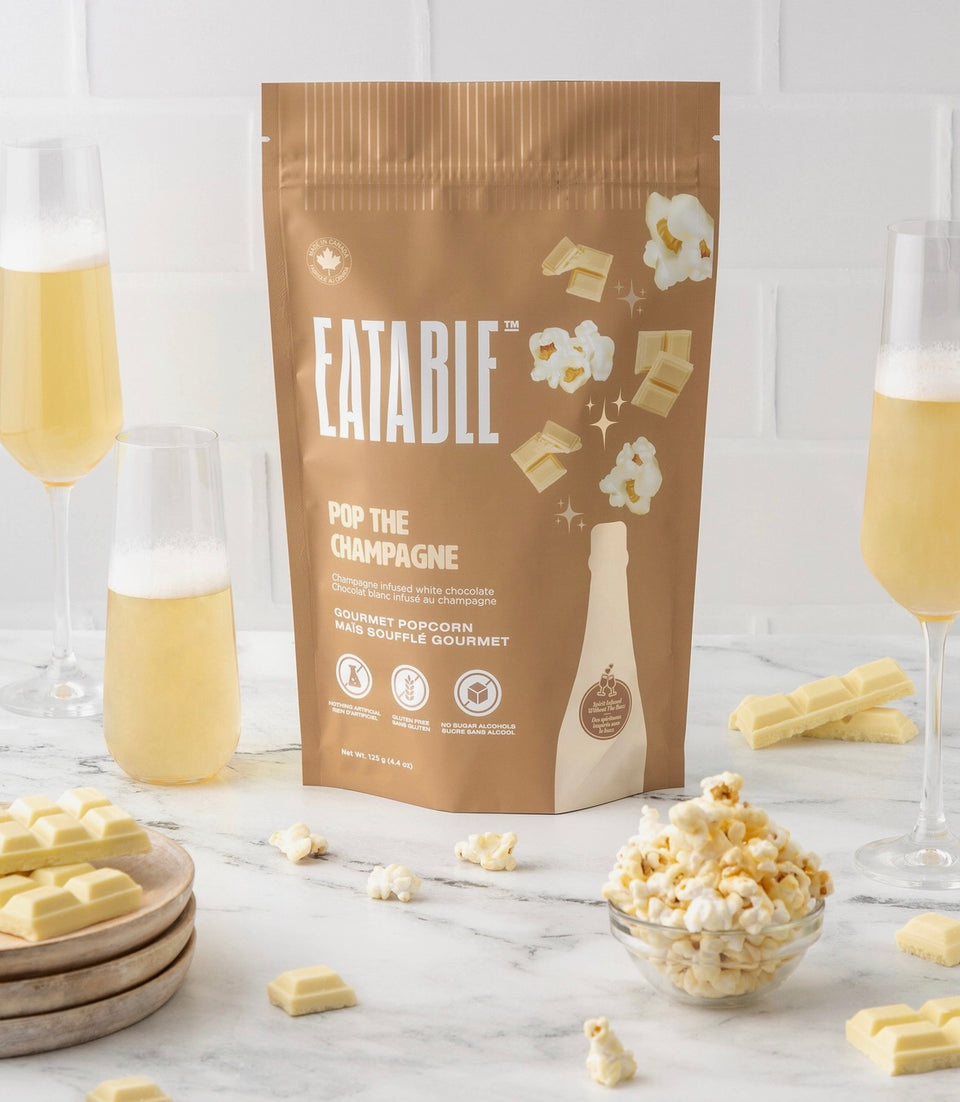 Package of champagne infused popcorn surrounded by popcorn, white chocolate pieces and glasses of Champagne 