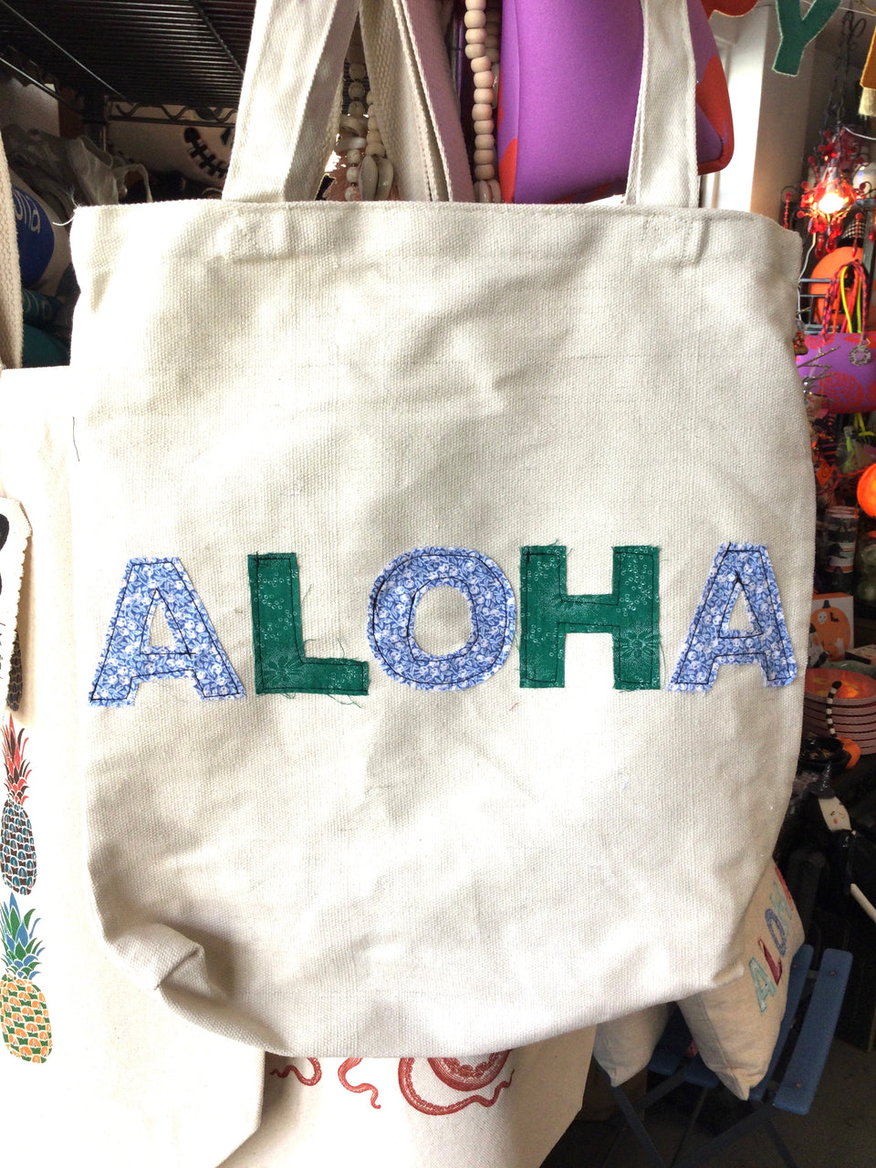 Cotton canvas tote with aloha is block letters cute out of bright colored and patterned fabric and hand stitched on the tote.