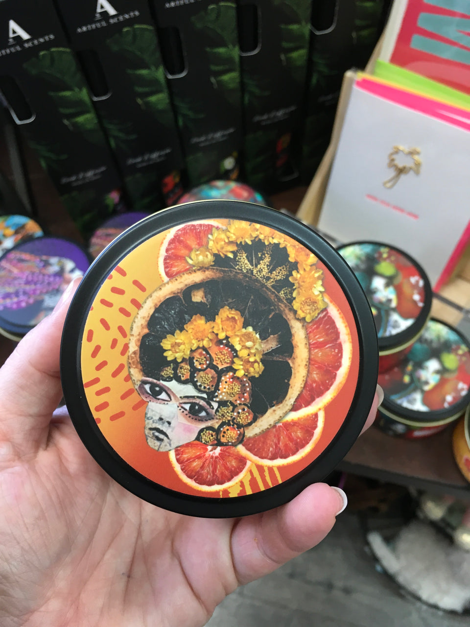 Top of round candle tin. Bold oranges and a dramatic face with big eyes and hair made of flowers.