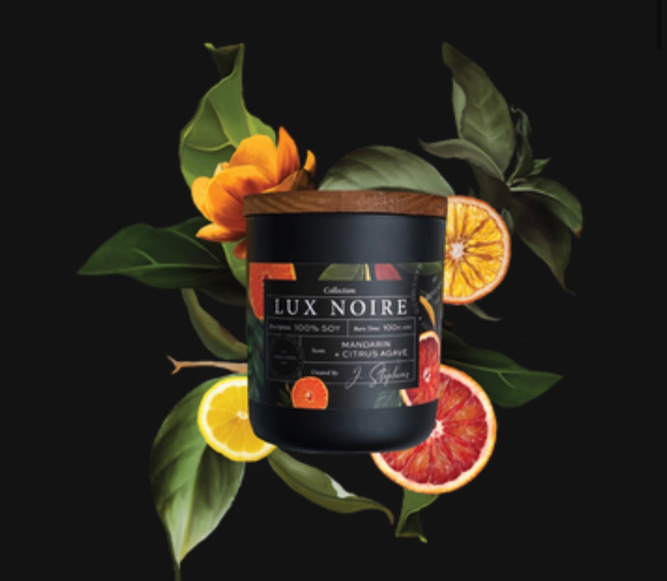 Lux Noire Candle - candle laying on the bed of cirtus fruits and leaves .Black frosted glass with wooden top and oranges and other cirus fruits printed on the label.