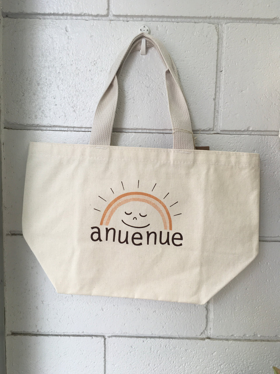 Canvas tote with Anuenue printed on the front which means rainbos in hawaiian. The rainbow has a smile face underneath it. bag is hanging on a white brick wall