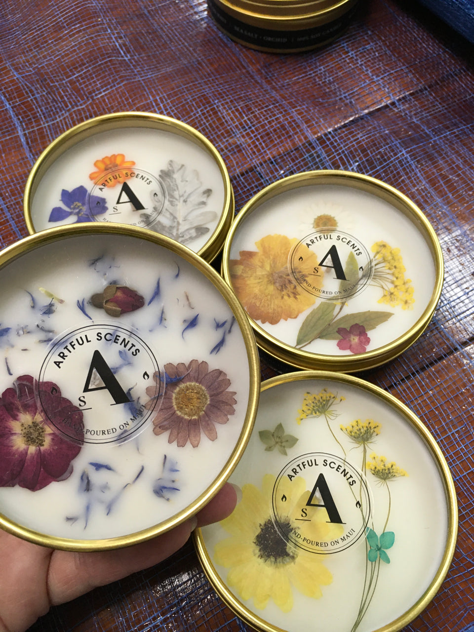 hand holding a candle with pressed flowers ont he top . logo is also visible A for Artful Scents. there are four different flower combinations in th photo